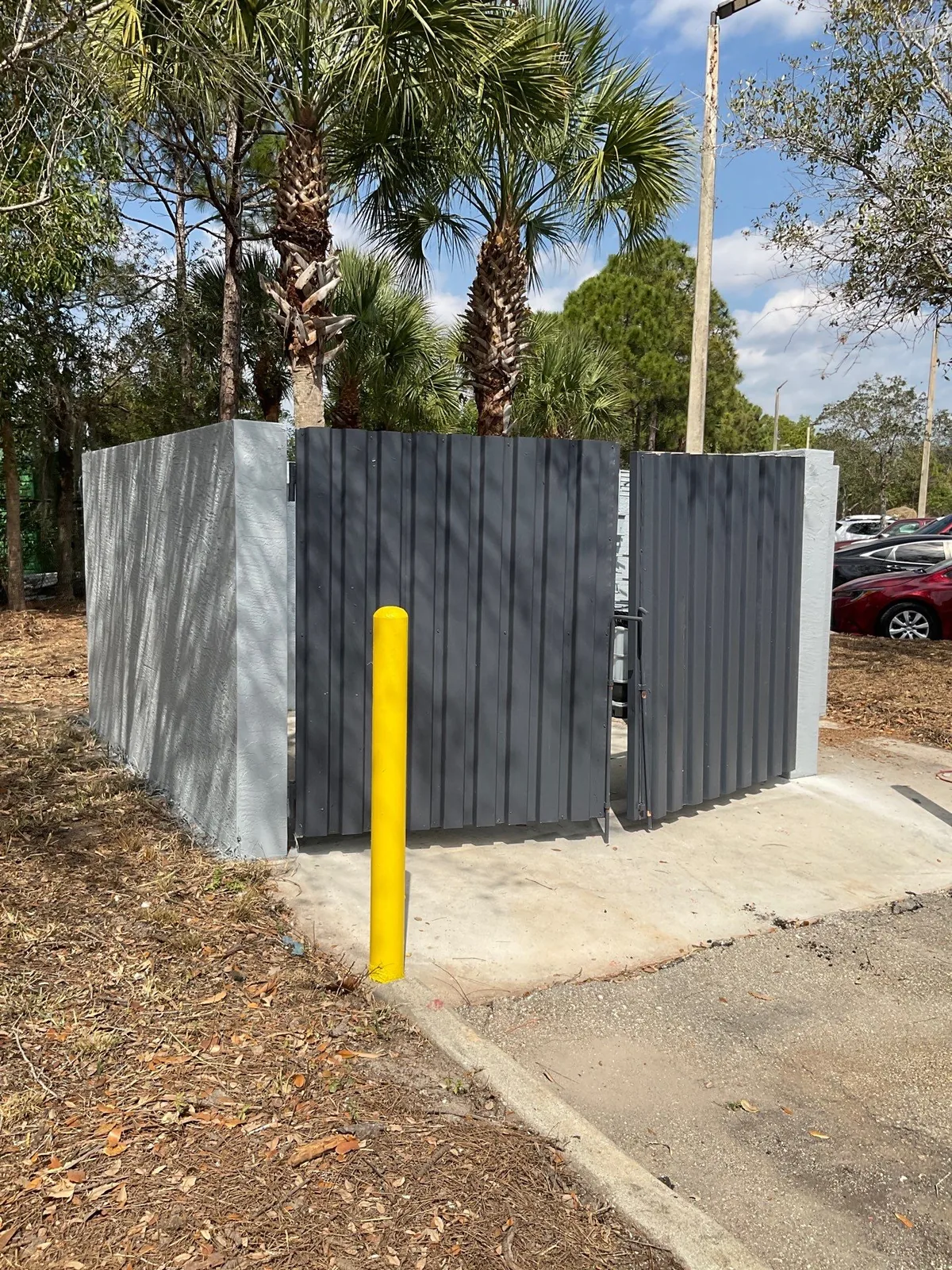 Commercial Painting in Jupiter, Florida. Painted grey dumpster enclosure with corrugated metal gates and block walls, yellow bollard in front, palms behind.