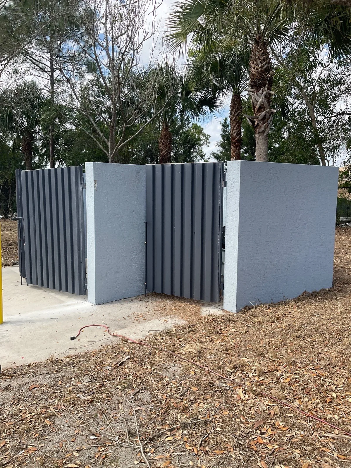 Commercial Painting in Jupiter, Florida. Freshly painted grey masonry dumpster enclosure with dark grey metal gates and white-capped pilasters on a concrete pad.