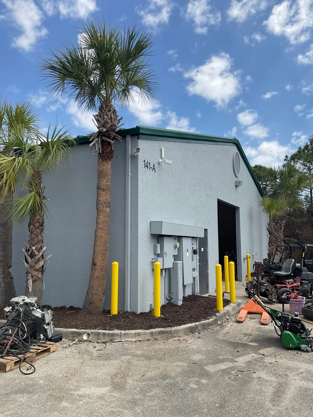 Commercial Painting in Jupiter, Florida. Single-storey commercial warehouse building with light grey painted walls, green roof trim and yellow bollards, palm trees in front.