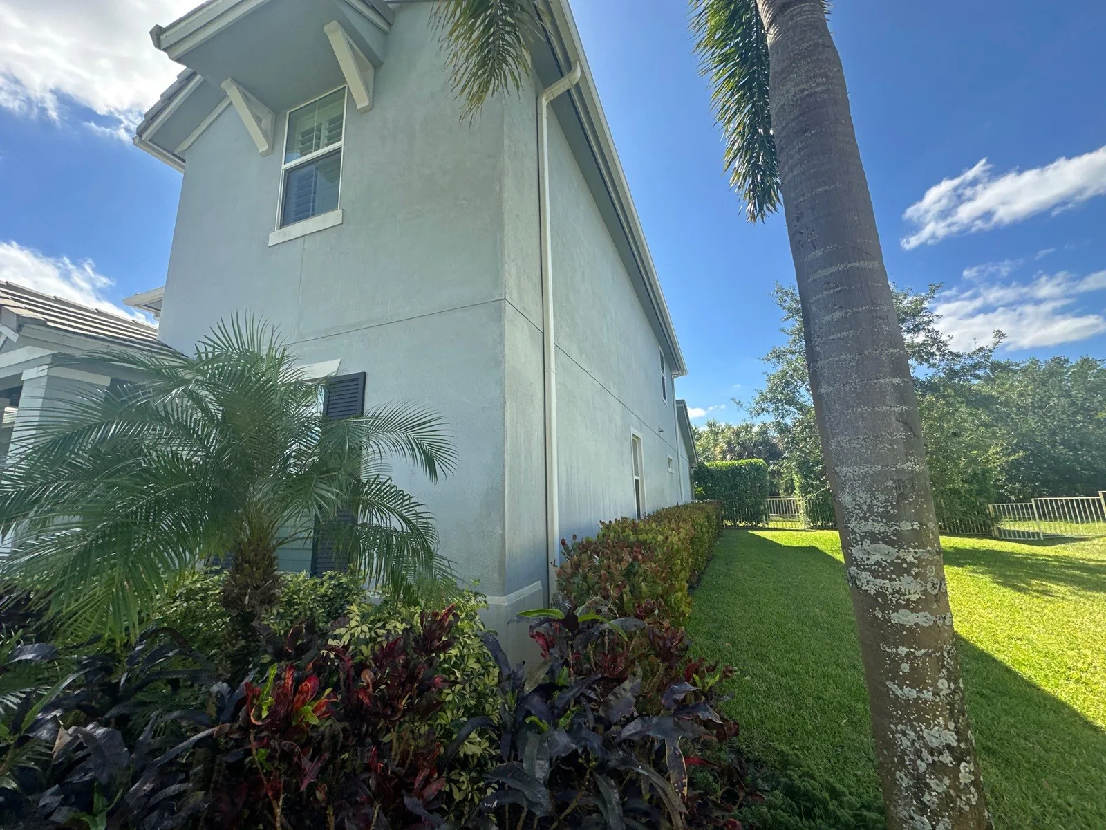 Exterior Painting in Jupiter, Florida. Side elevation of a two-storey pale blue-grey stucco house with white trim and dark shutters, framed by a palm and landscaping under a blue sky.