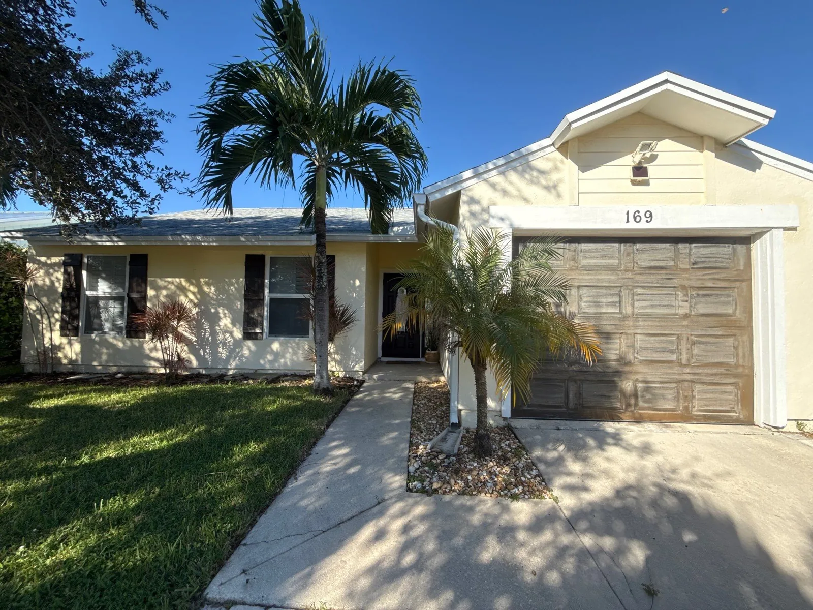 Exterior Painting in Jupiter, Florida. Single-storey home exterior, pale cream stucco with white trim, dark shutters, brown garage door and palm tree in the front yard.
