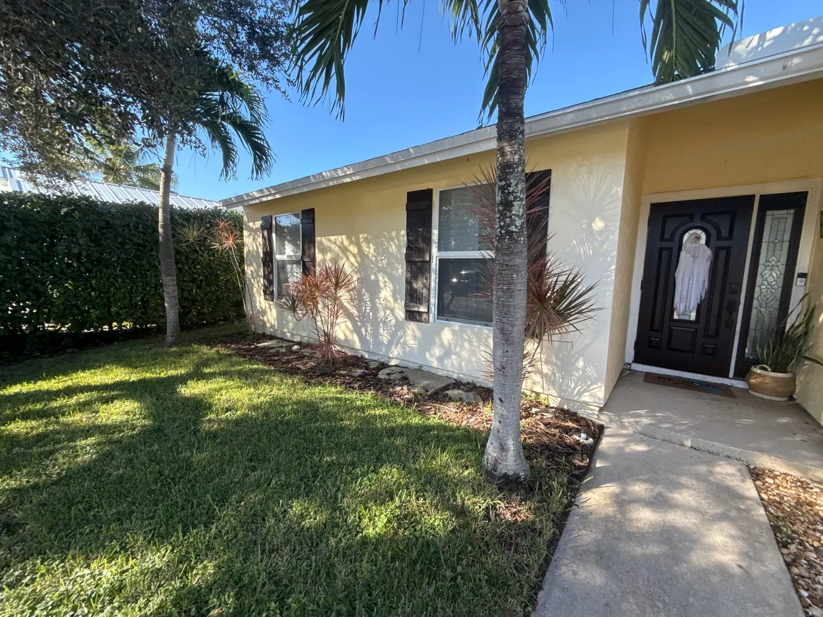 Exterior Painting in Jupiter, Florida. Front elevation of a single-storey home, yellow stucco with white fascia, dark front door and palm trees over a lawn.