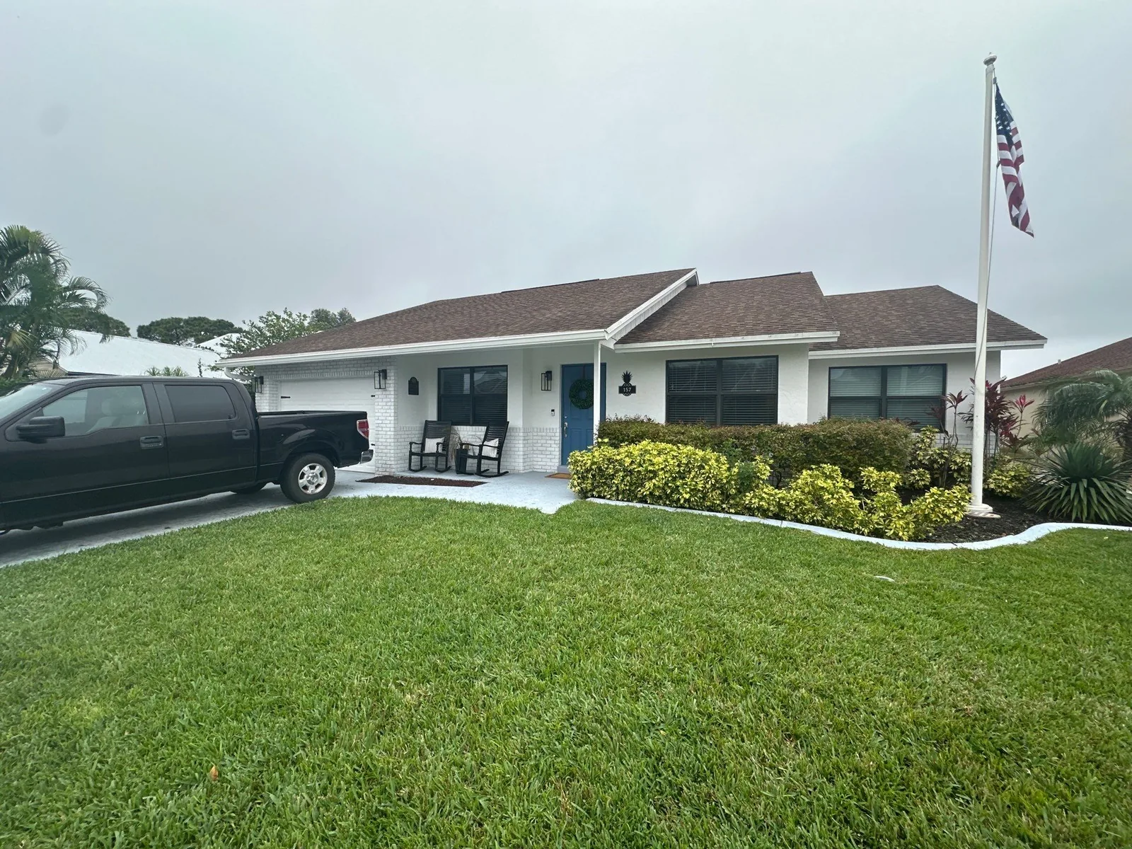 Exterior Painting in Jupiter, Florida. Single-storey home painted white with white trim, a blue front door and brown shingle roof, clipped hedges and lawn, with a pickup truck parked in the driveway.