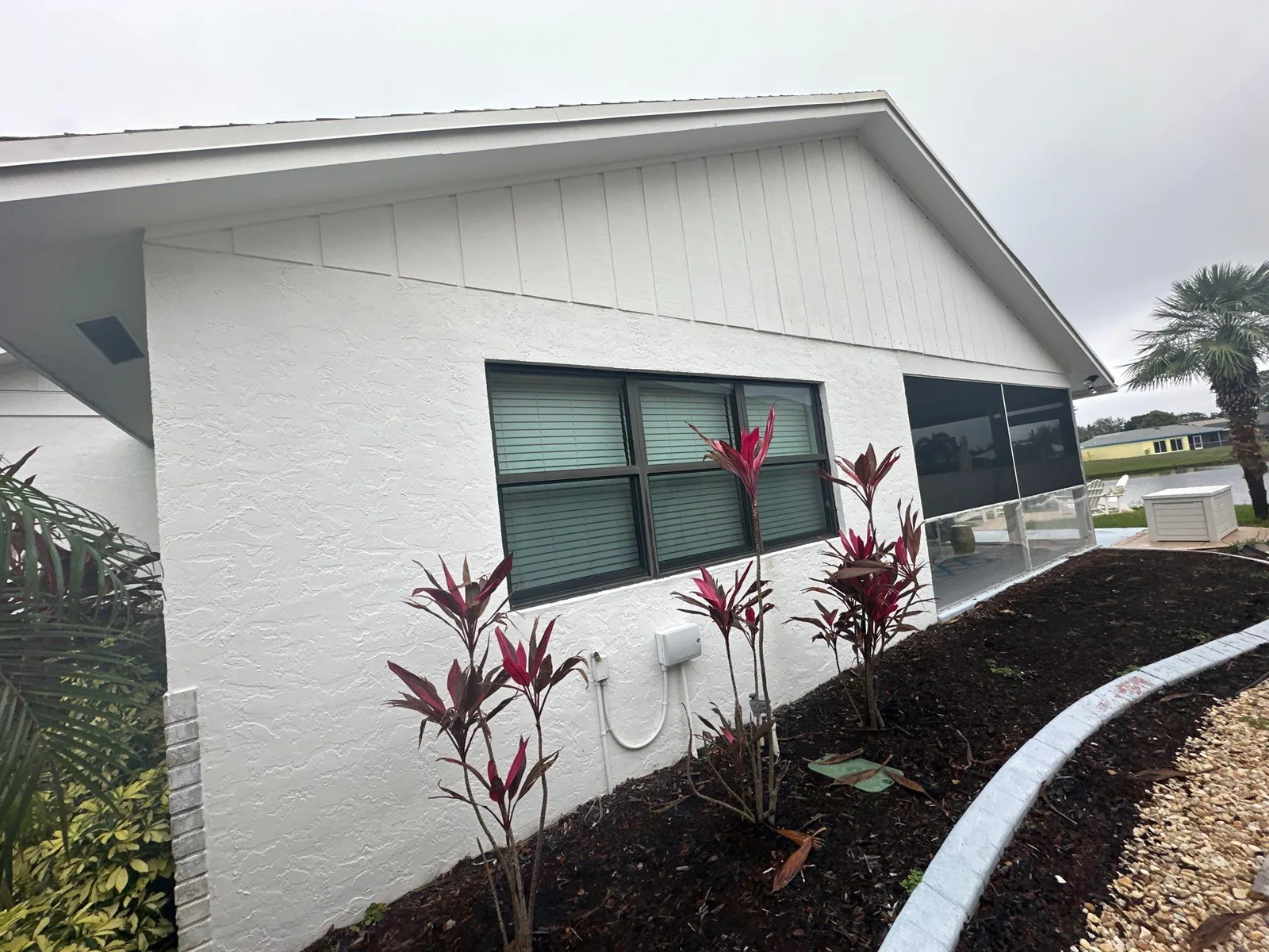Exterior Painting in Jupiter, Florida. Single-storey house with white painted stucco and board-and-batten gable, dark-framed windows and a planted mulch bed.