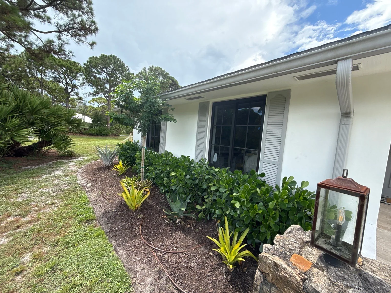 Exterior Painting in Jupiter, Florida. White stucco single-storey home exterior with white shutters, dark window and landscaped shrub bed.