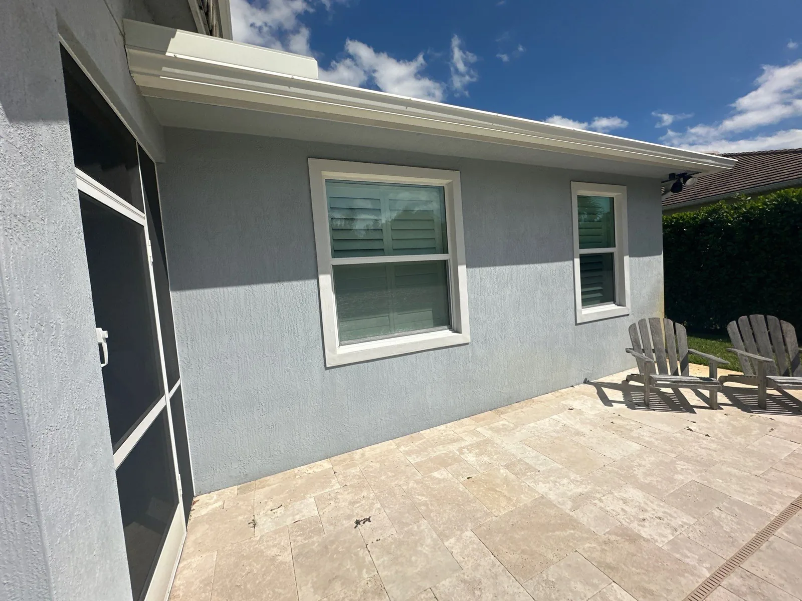 Exterior Painting in Jupiter, Florida. Exterior stucco wall painted blue-grey with white window trim and soffit, a screened door at left and a travertine paver patio.