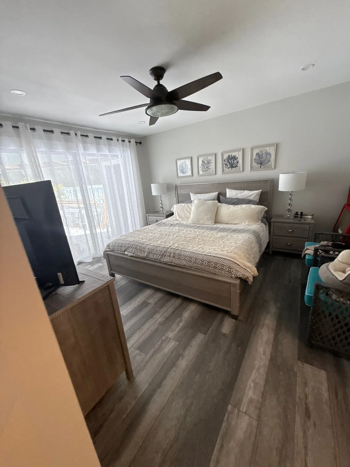 Interior Painting in Jupiter, Florida. Bedroom with light grey walls and white ceiling, made bed with upholstered headboard, framed botanical prints, matching lamps and grey wood-look flooring.