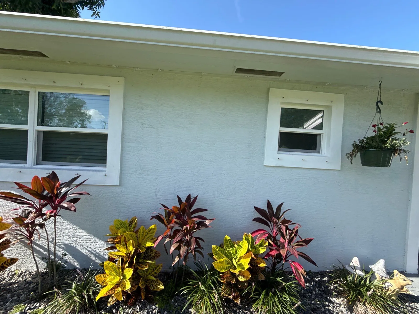 Exterior Painting in Lakewood Park, Florida. Pale grey-white stucco exterior wall with two white-trimmed windows, a hanging flower basket and a bed of crotons in front.