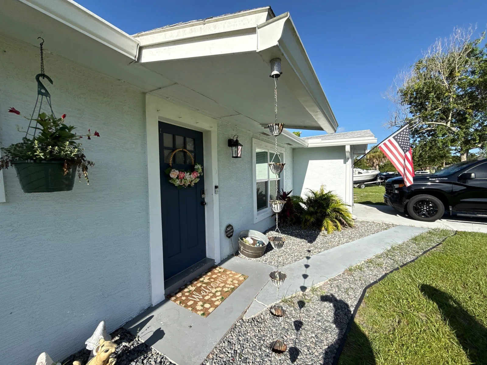 Exterior Painting in Lakewood Park, Florida. Front entry of a single-storey house with pale off-white walls, a dark navy front door with a wreath, hanging flower baskets, a gravel bed and a dark SUV parked at right.