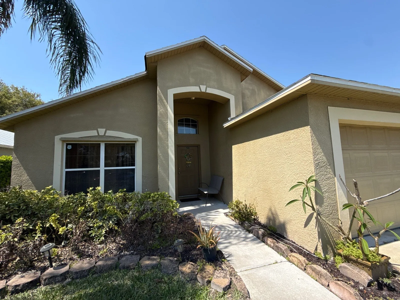 Exterior Painting in Melbourne, Florida. Single-storey house front elevation, tan stucco with white trim, arched entry and attached garage.