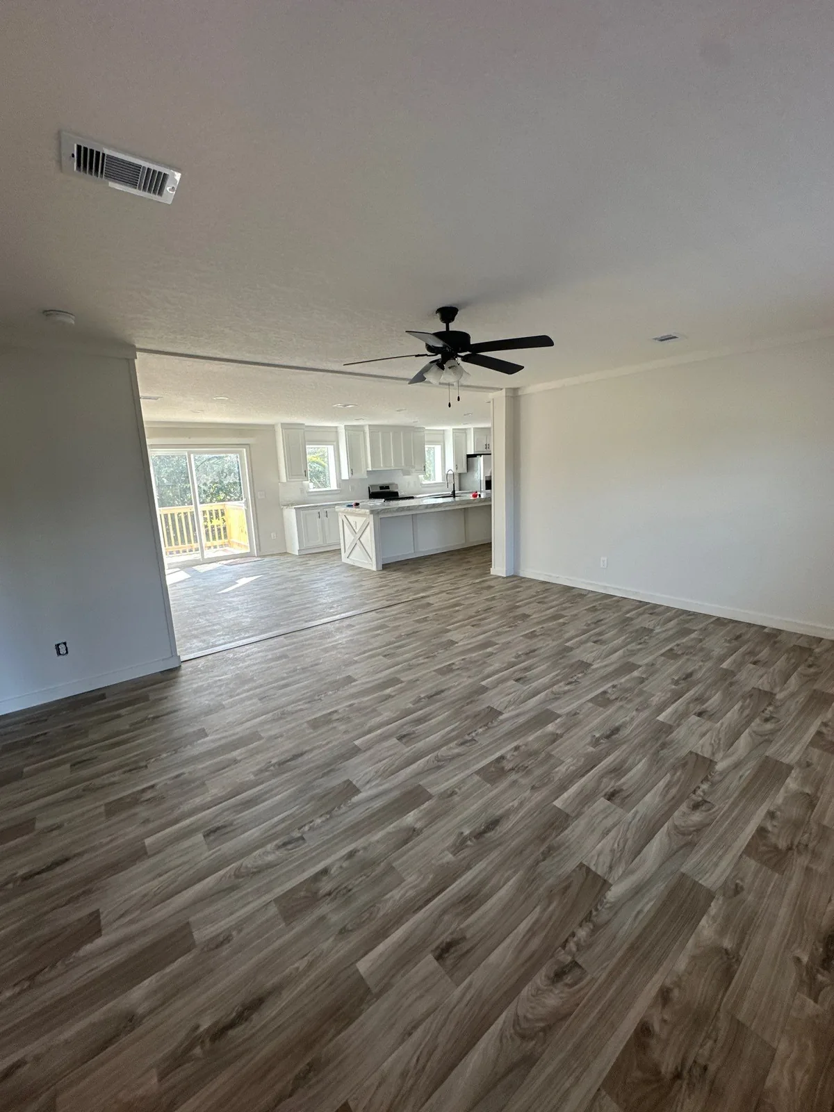 Interior Painting in Melbourne, Florida. Empty open-plan living room with white walls and ceiling, crown moulding, ceiling fan and wood-look plank flooring, kitchen visible beyond.