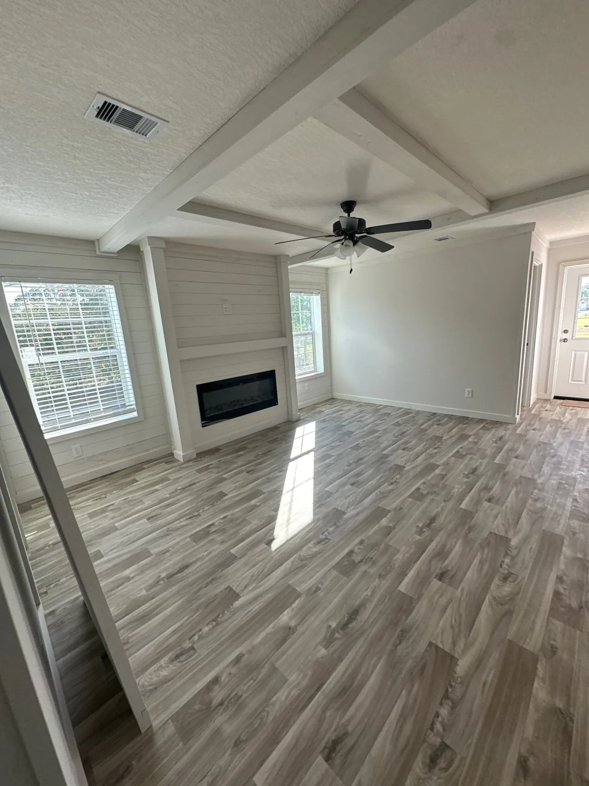 Interior Painting in Melbourne, Florida. Empty living room with a white shiplap fireplace wall, painted ceiling beams, white walls, ceiling fan and wood-look plank flooring.