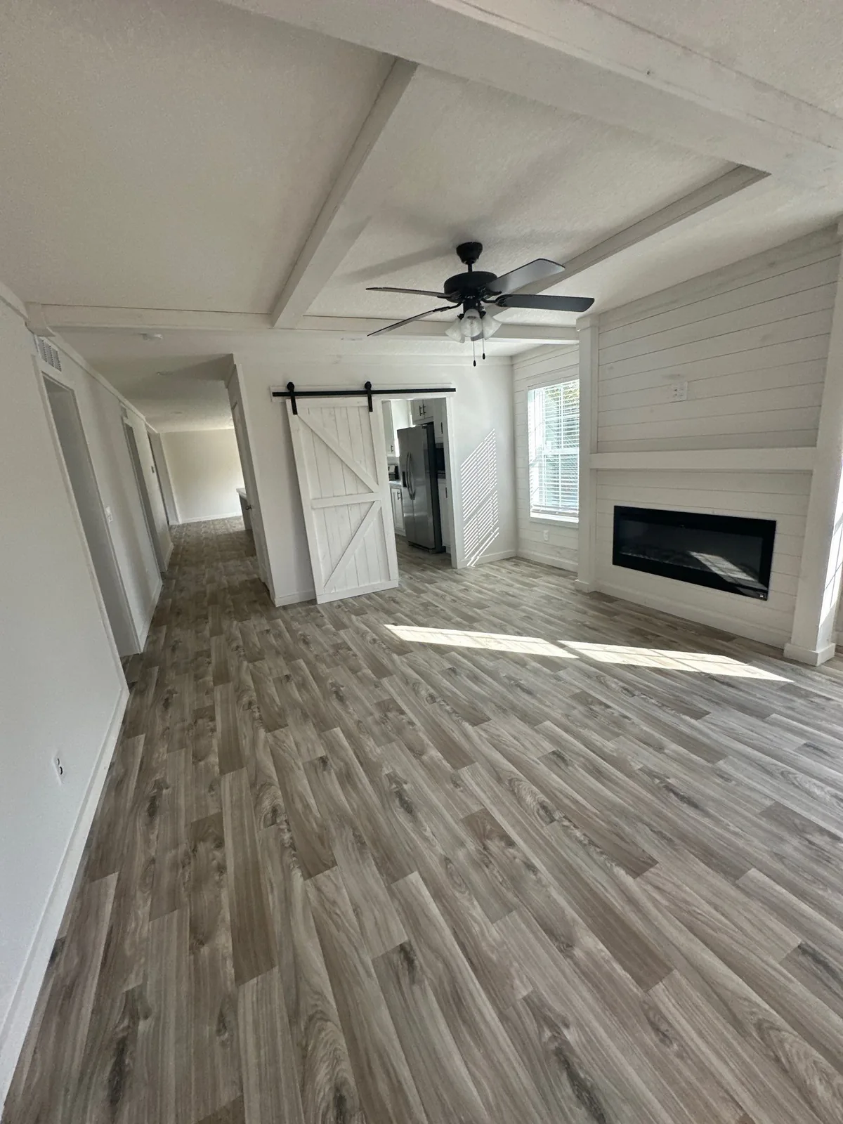 Interior Painting in Melbourne, Florida. Empty open-plan living room with white walls and beamed ceiling, white shiplap fireplace wall, sliding barn door and wood-look plank flooring.