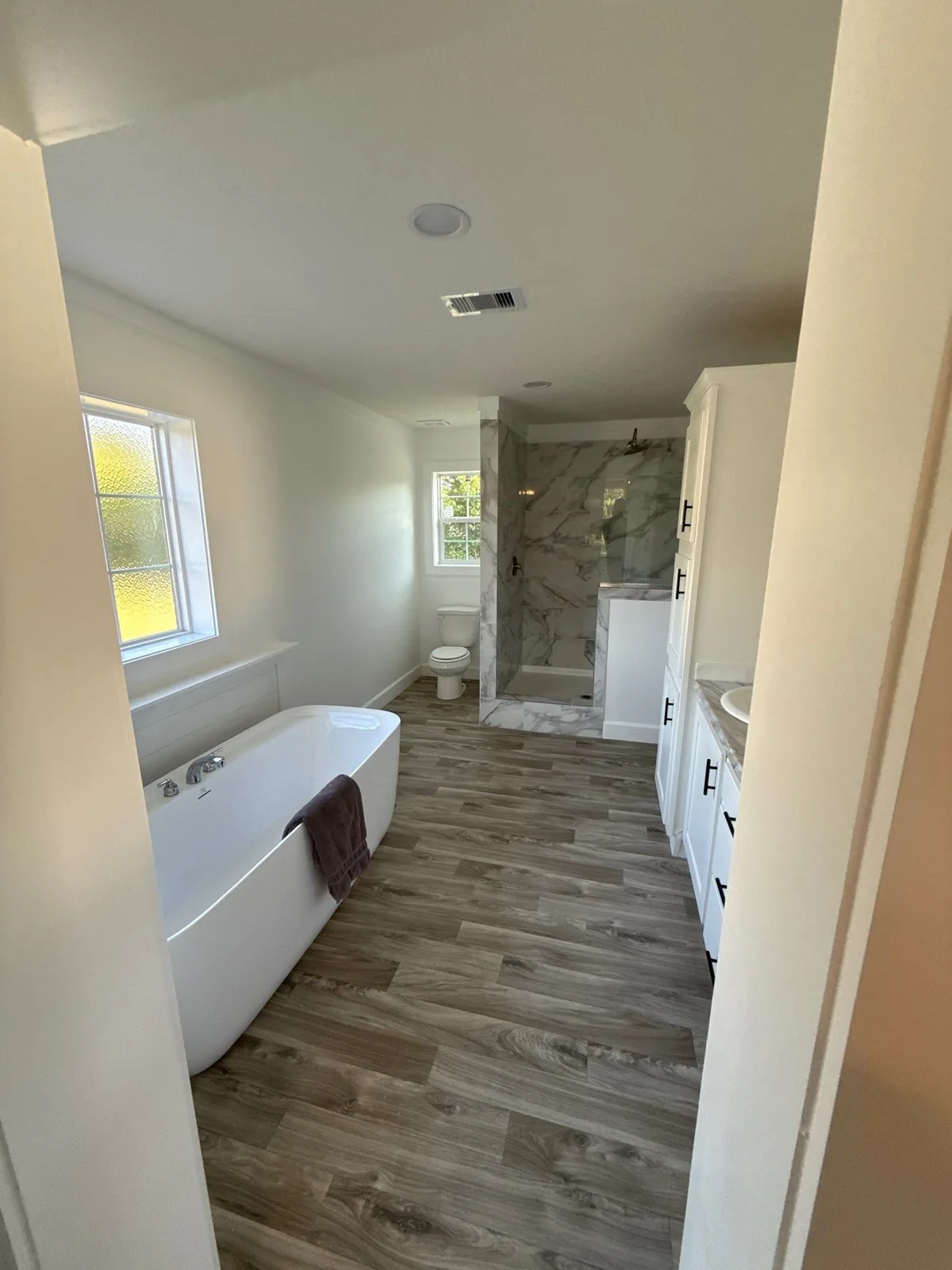 Interior Painting in Melbourne, Florida. Bathroom with white painted walls, a freestanding white soaking tub under a window, a tiled walk-in shower with glass panel, and wood-look plank flooring.