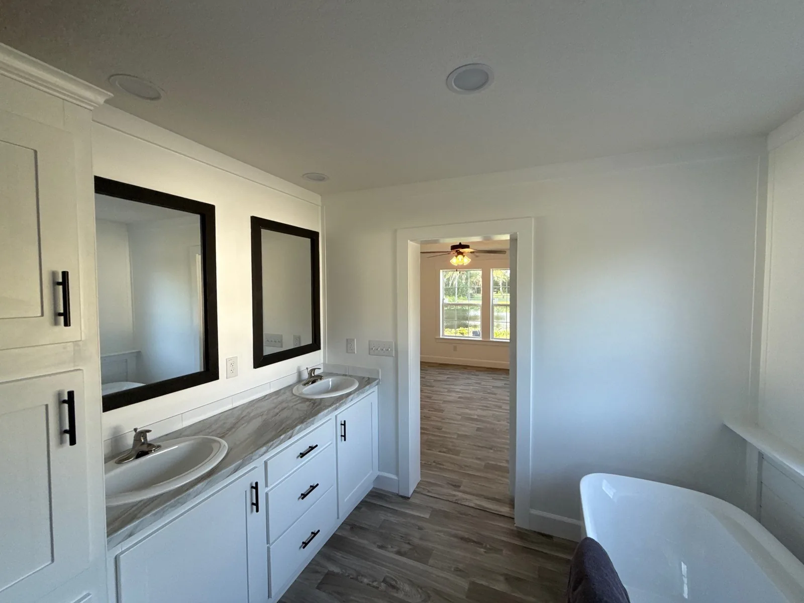 Interior Painting in Melbourne, Florida. Bathroom with a white double vanity, two black-framed mirrors, white painted walls and trim, and a view through the doorway into a bedroom.