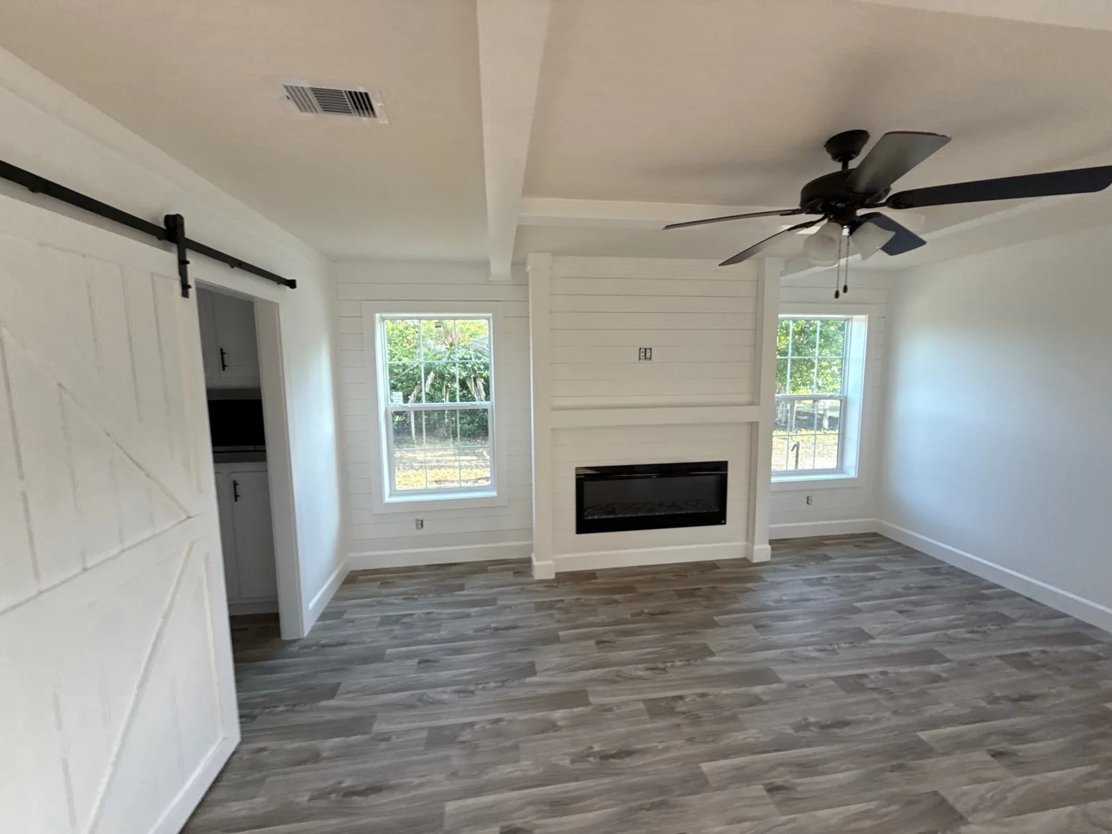 Interior Painting in Melbourne, Florida. Empty living room with white shiplap fireplace surround, white walls and trim, beamed ceiling, ceiling fan and a white sliding barn door.