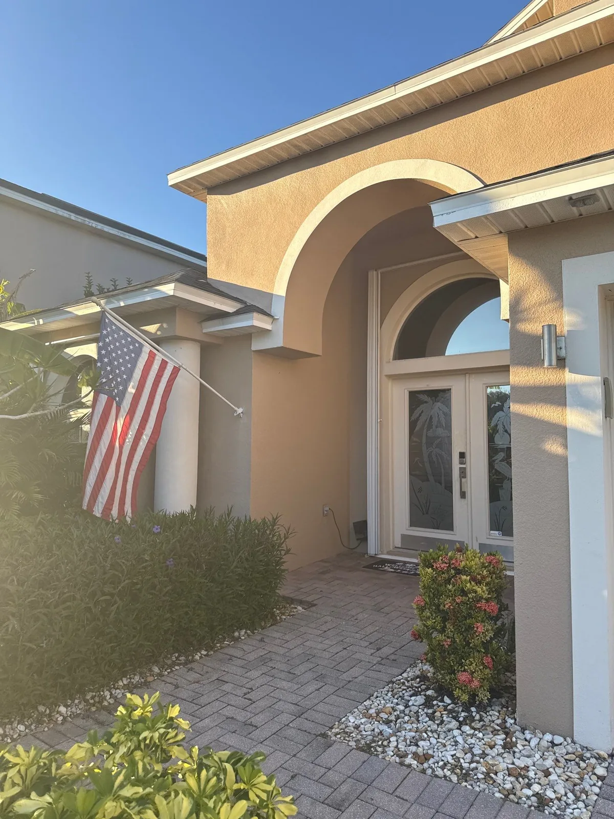 Exterior Painting in Merritt Island, Florida. Arched front entry with tan stucco, white trim and glass double doors, flag and paver walkway in evening light.