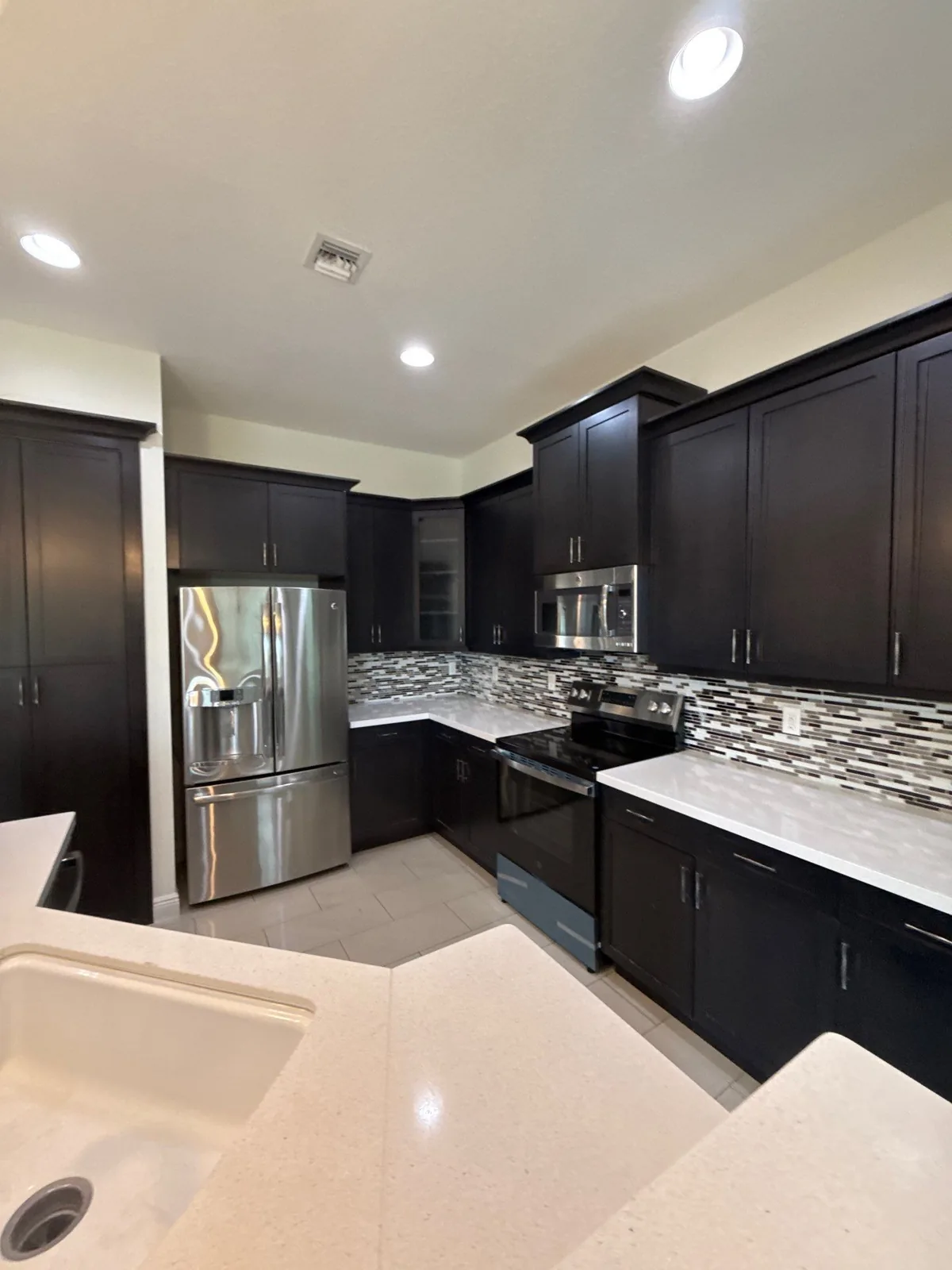 Cabinet Refinishing in North Palm Beach, Florida. Kitchen with dark brown cabinets, stainless steel appliances, mosaic tile backsplash and light quartz countertops.