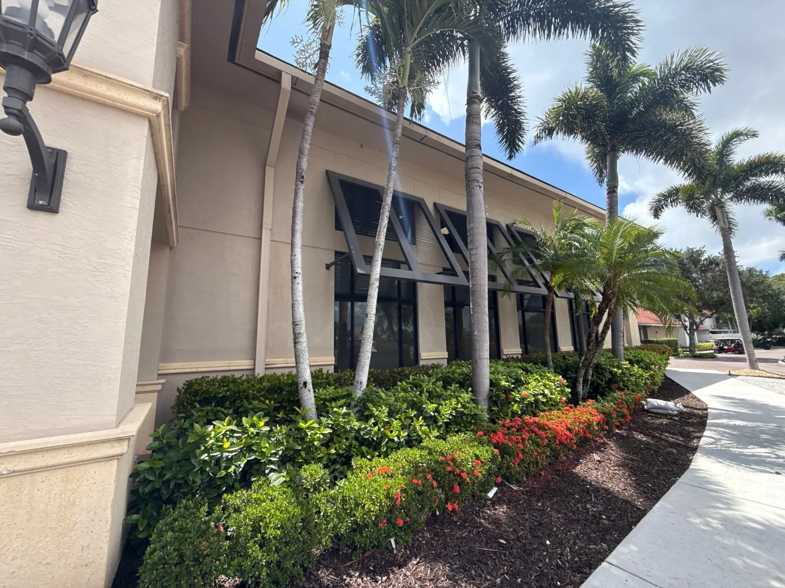 Commercial Painting in North Palm Beach, Florida. Single-storey commercial building with beige stucco walls, dark bahama shutters over the windows and palms and flowering hedge in front.
