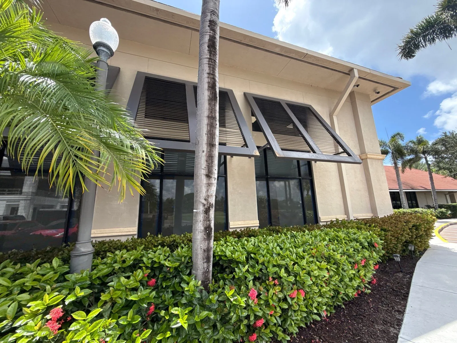 Commercial Painting in North Palm Beach, Florida. Beige stucco commercial facade with dark bahama shutters and dark storefront glazing, hedge and lamp post in front.