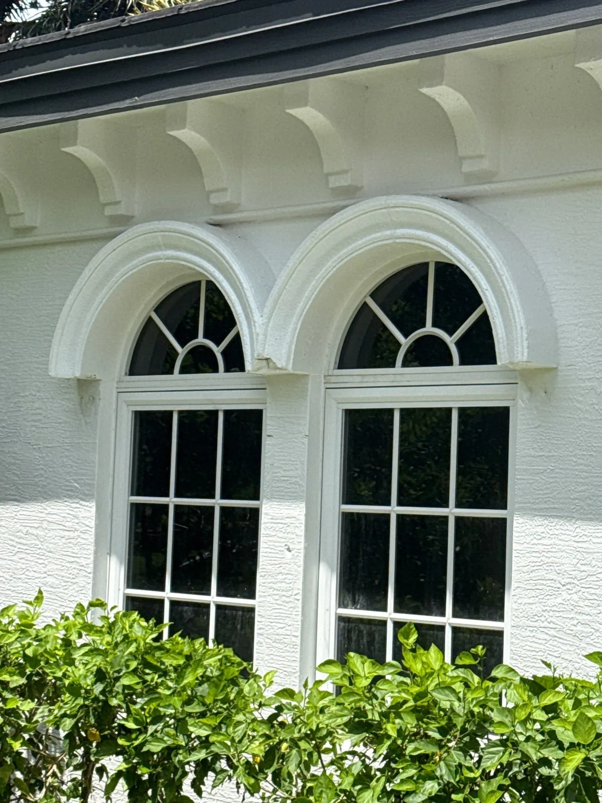 Exterior Painting in North Palm Beach, Florida. White stucco exterior with paired arched multi-pane windows and scalloped corbels beneath a dark eave, hedge in the foreground.