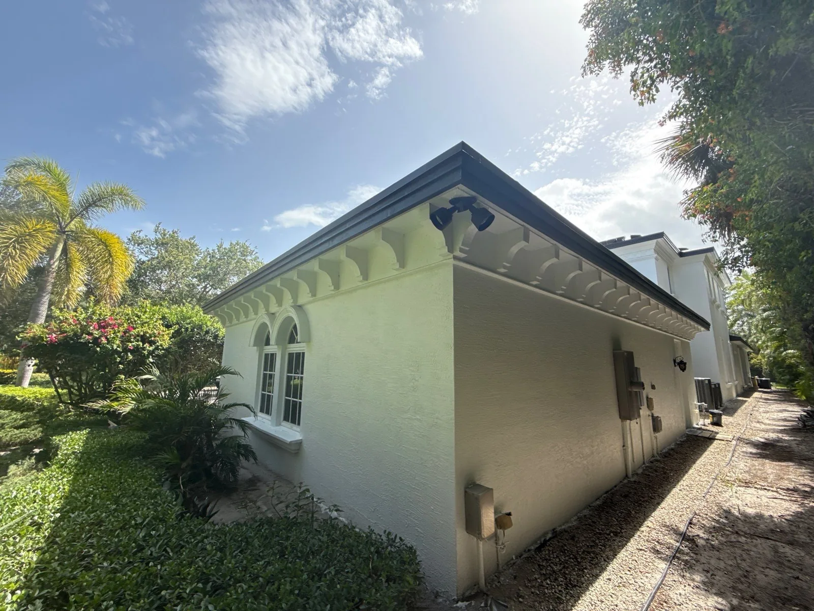 Exterior Painting in North Palm Beach, Florida. Corner of a cream stucco single-storey house with dark fascia, decorative eave corbels and an arched window.