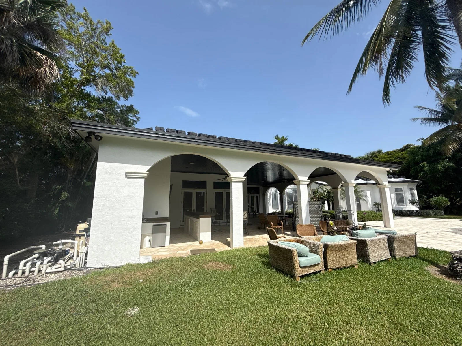 Exterior Painting in North Palm Beach, Florida. Rear elevation of a white stucco house with arched covered patio, dark fascia and wicker seating on the lawn.
