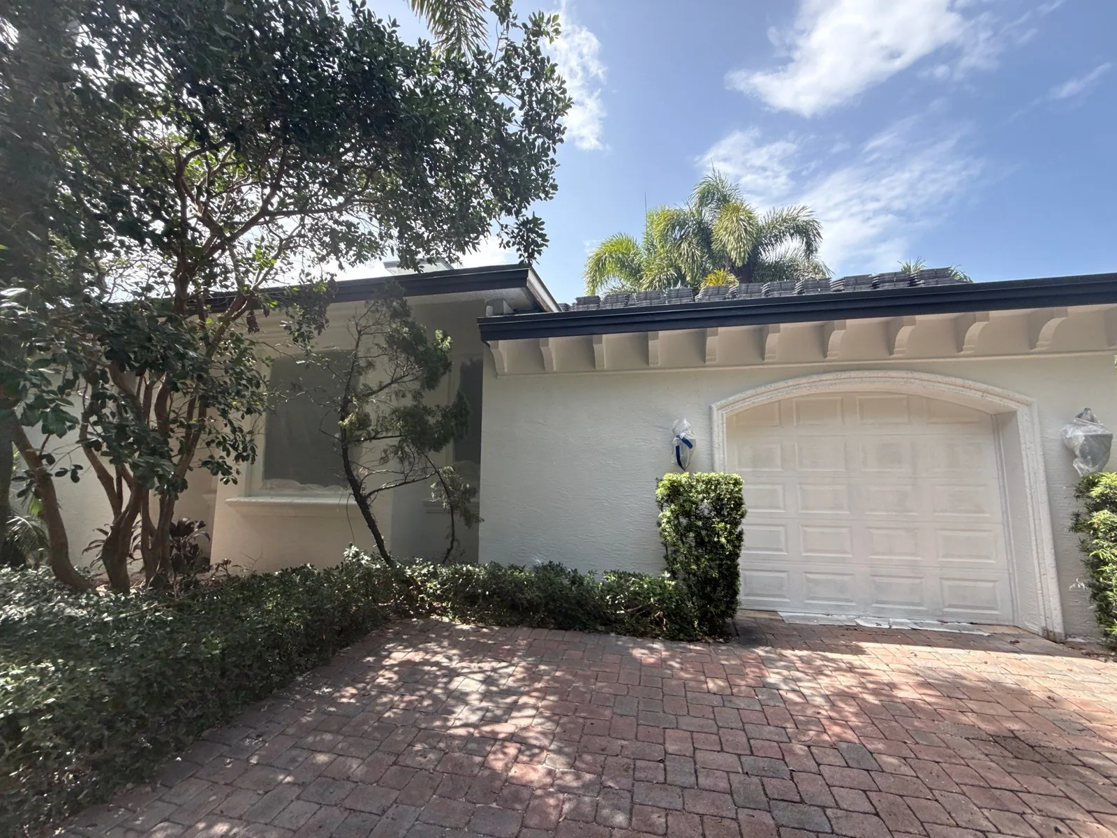 Exterior Painting in North Palm Beach, Florida. Front elevation of a single-storey home with light stucco walls, an arched white garage door, trimmed hedges and a brick paver driveway.