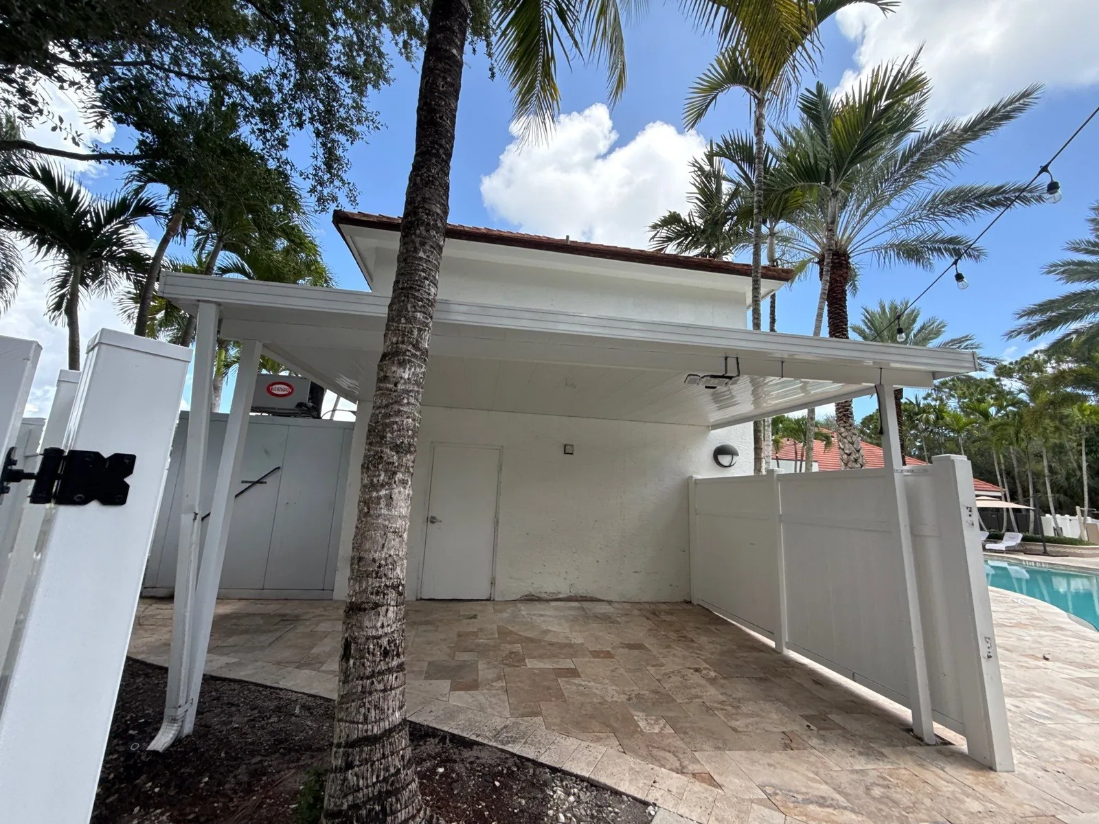 Exterior Painting in North Palm Beach, Florida. White painted poolside cabana with a flat overhang, travertine paving and white vinyl fencing beside a pool.