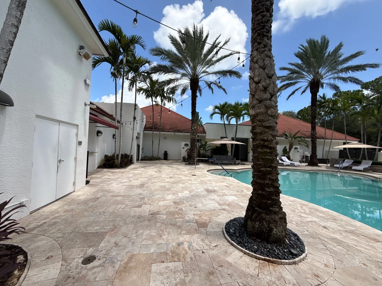 Exterior Painting in North Palm Beach, Florida. White painted stucco pool house wall beside a travertine pool deck, swimming pool and palms under blue sky.