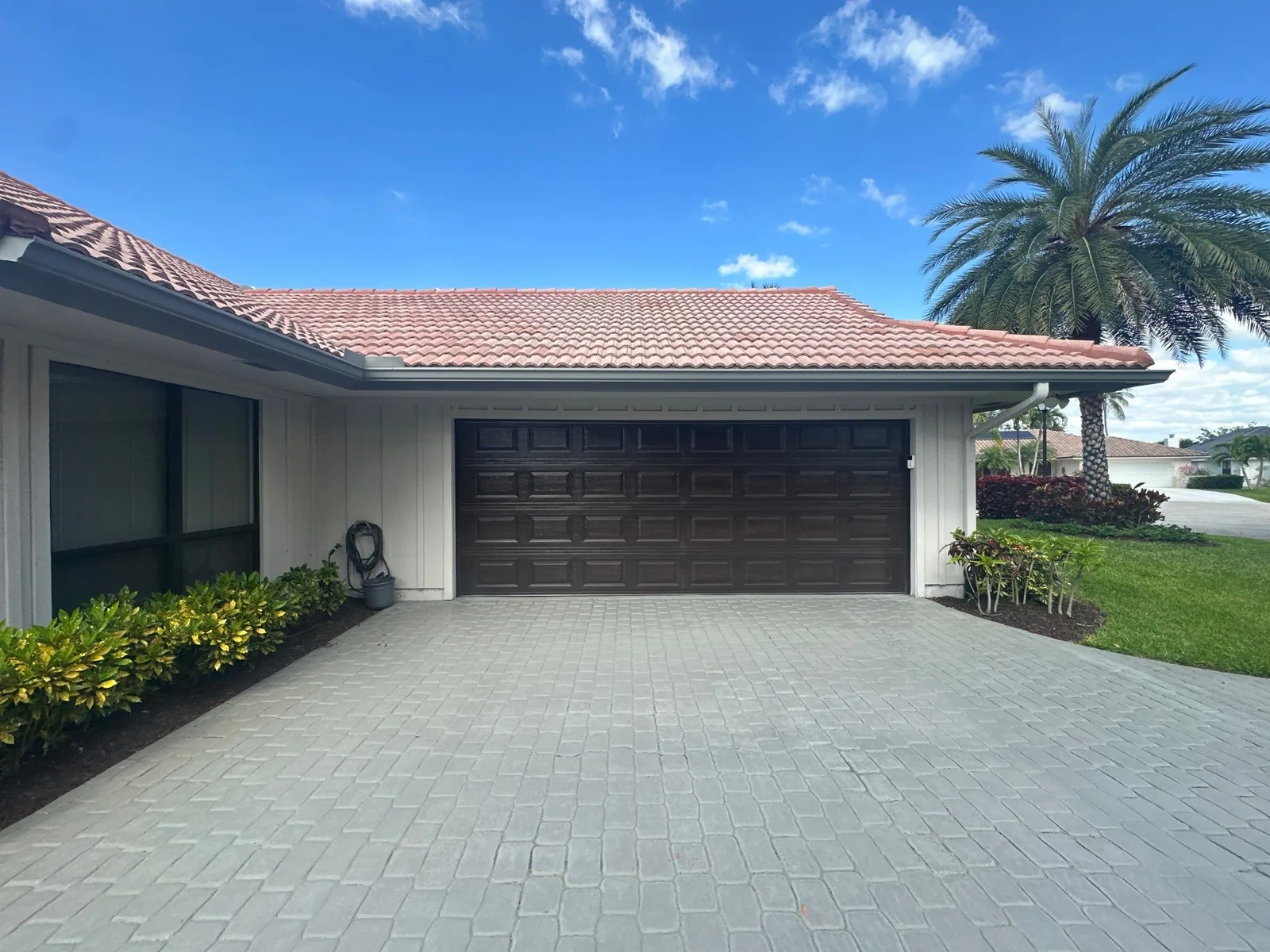 Exterior Painting in North Palm Beach, Florida. Single-storey house with white board-and-batten siding, a dark brown garage door and a barrel-tile roof, seen from a paver driveway.