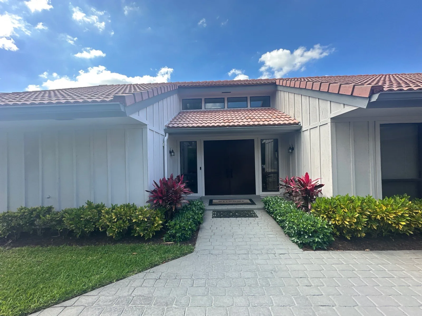 Exterior Painting in North Palm Beach, Florida. Front entry of a single-storey white-painted house with barrel-tile roof, trimmed hedges and a paver walkway under a blue sky.