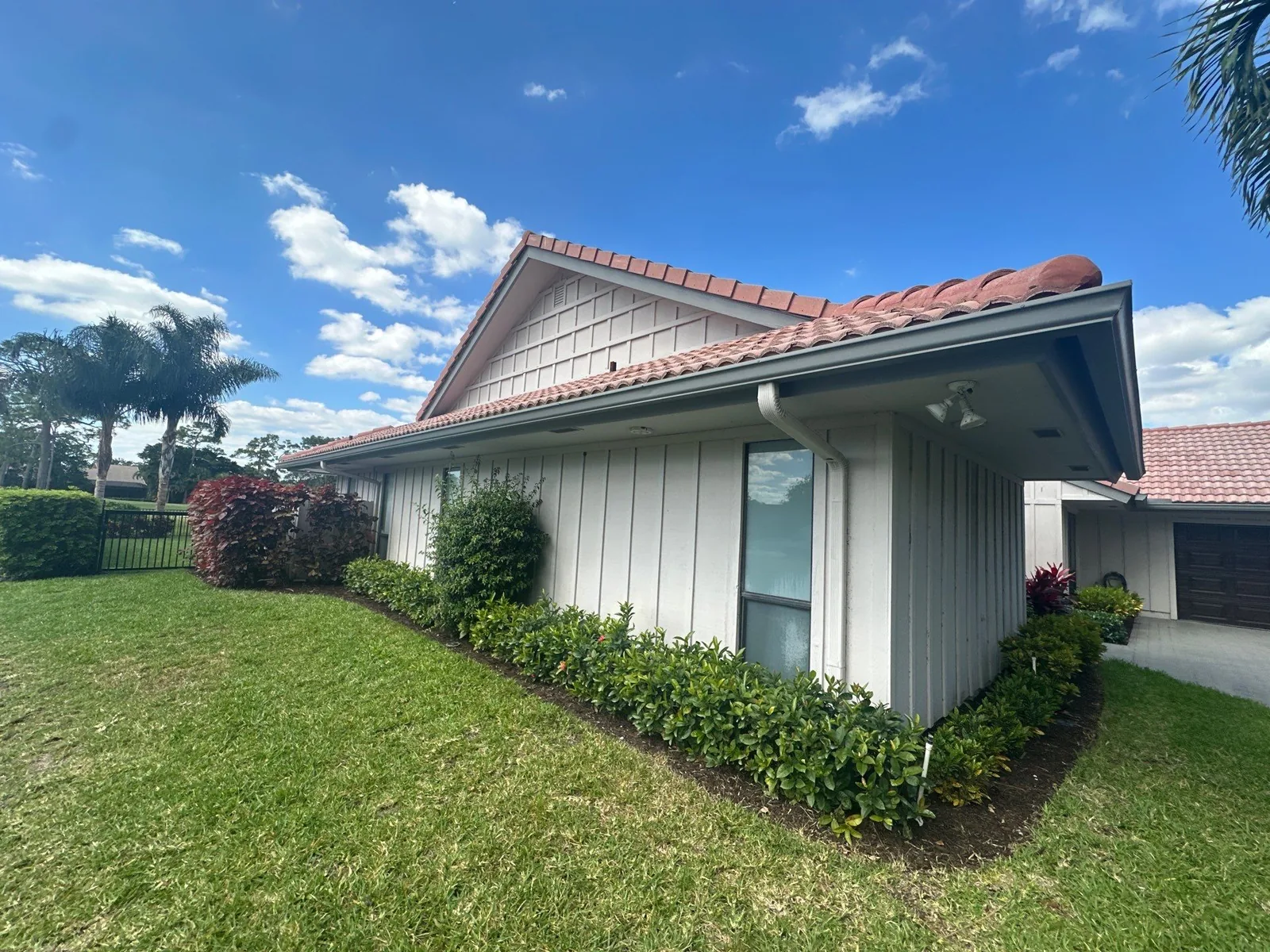 Exterior Painting in North Palm Beach, Florida. Gable end of a single-storey house with white board-and-batten siding and barrel-tile roof, with a hedge row along the base.
