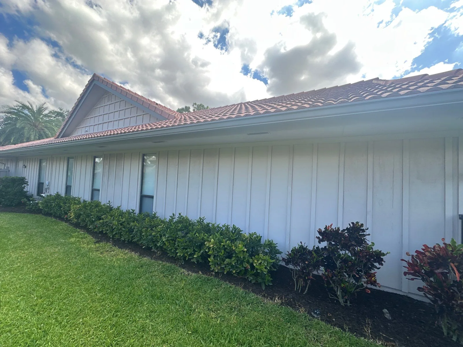 Exterior Painting in North Palm Beach, Florida. Long side wall of a single-storey house in white board-and-batten siding with barrel-tile roof and shrubs along the base.