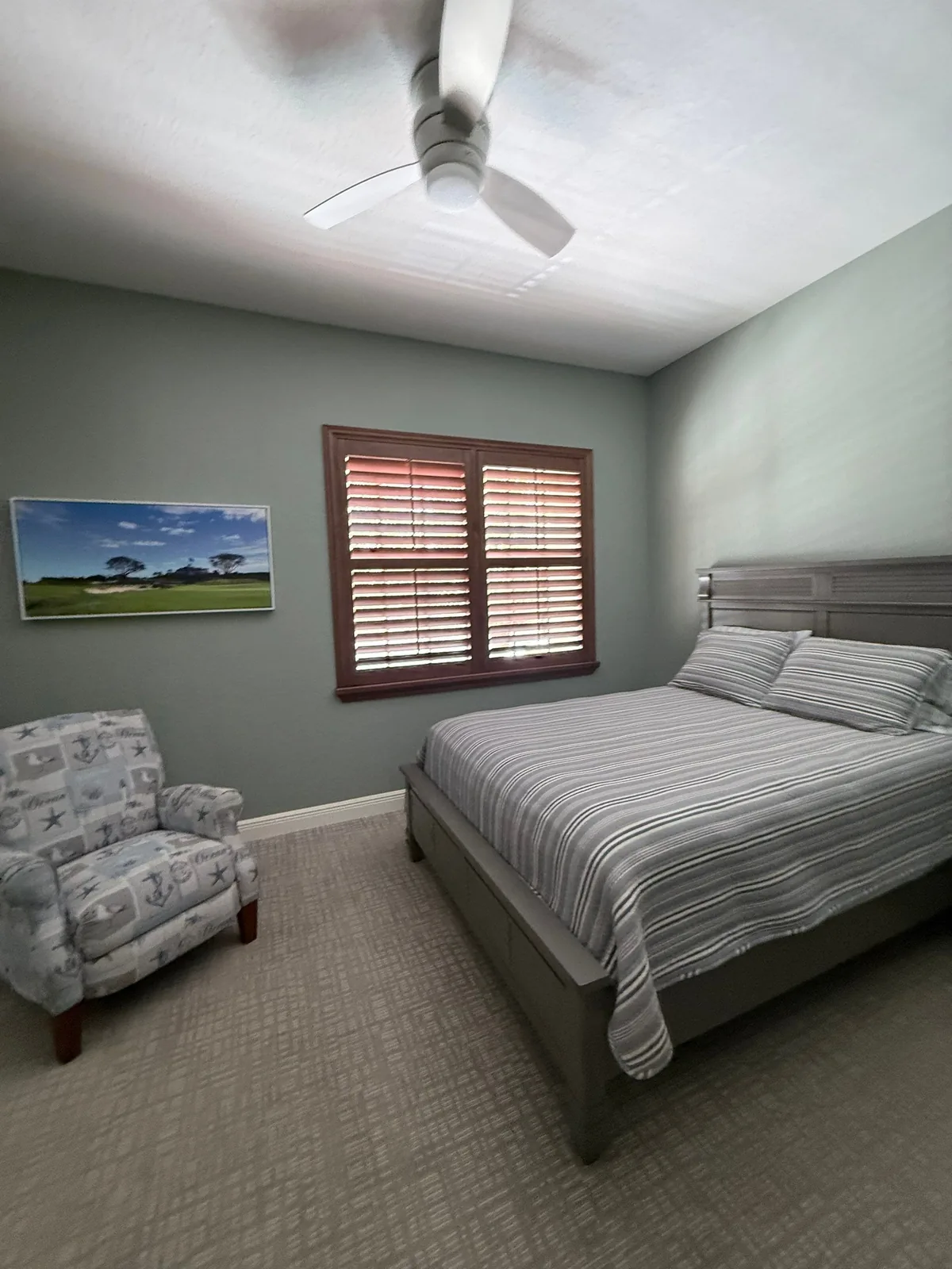 Interior Painting in North Palm Beach, Florida. Furnished bedroom with sage-green painted walls, white ceiling, dark wood window shutters and an upholstered armchair.