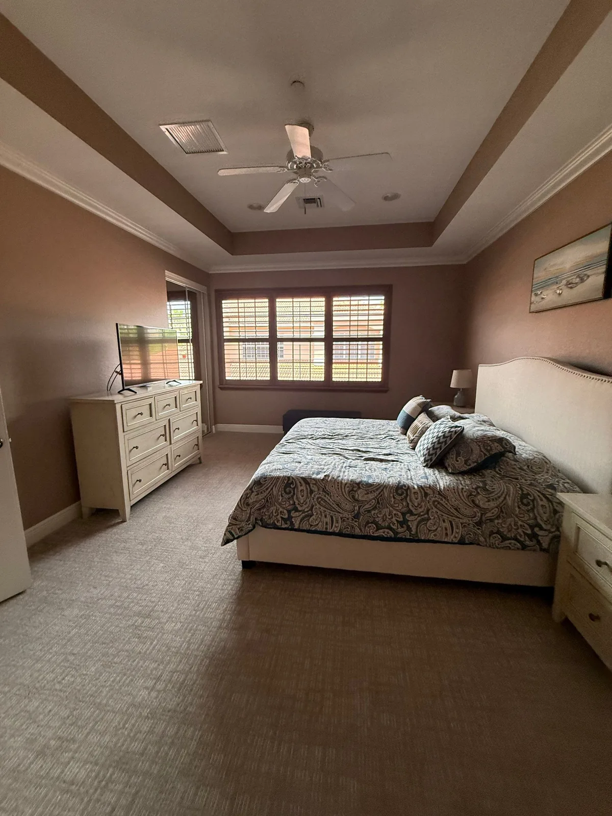 Interior Painting in North Palm Beach, Florida. Furnished bedroom with taupe walls, tray ceiling and ceiling fan, plantation shutters and a made bed with dresser.