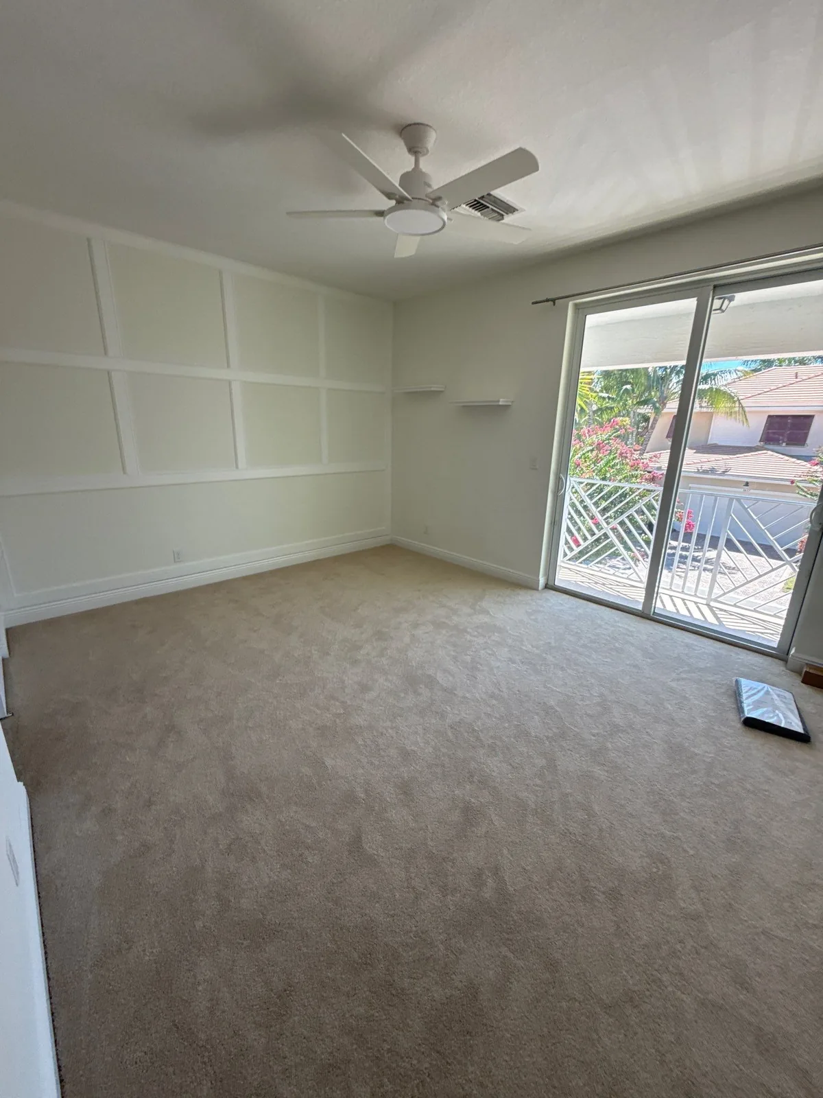 Interior Painting in North Palm Beach, Florida. Empty bedroom with a white picture-frame moulding accent wall, carpet, ceiling fan and sliding glass door to a balcony.