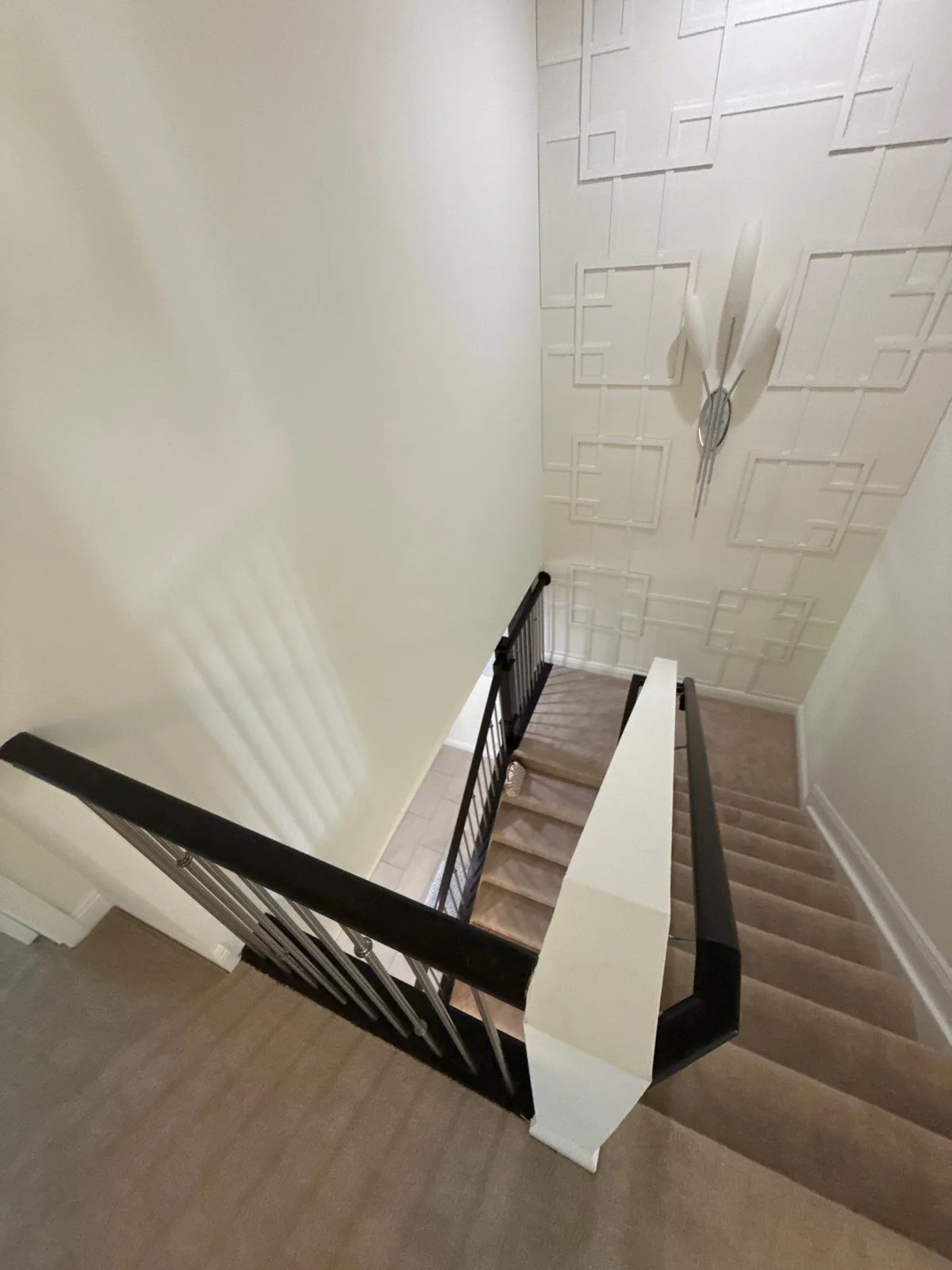 Interior Painting in North Palm Beach, Florida. Stairwell viewed from the landing above, with a white geometric raised-panel feature wall, dark painted handrail and newel, and carpeted stairs.