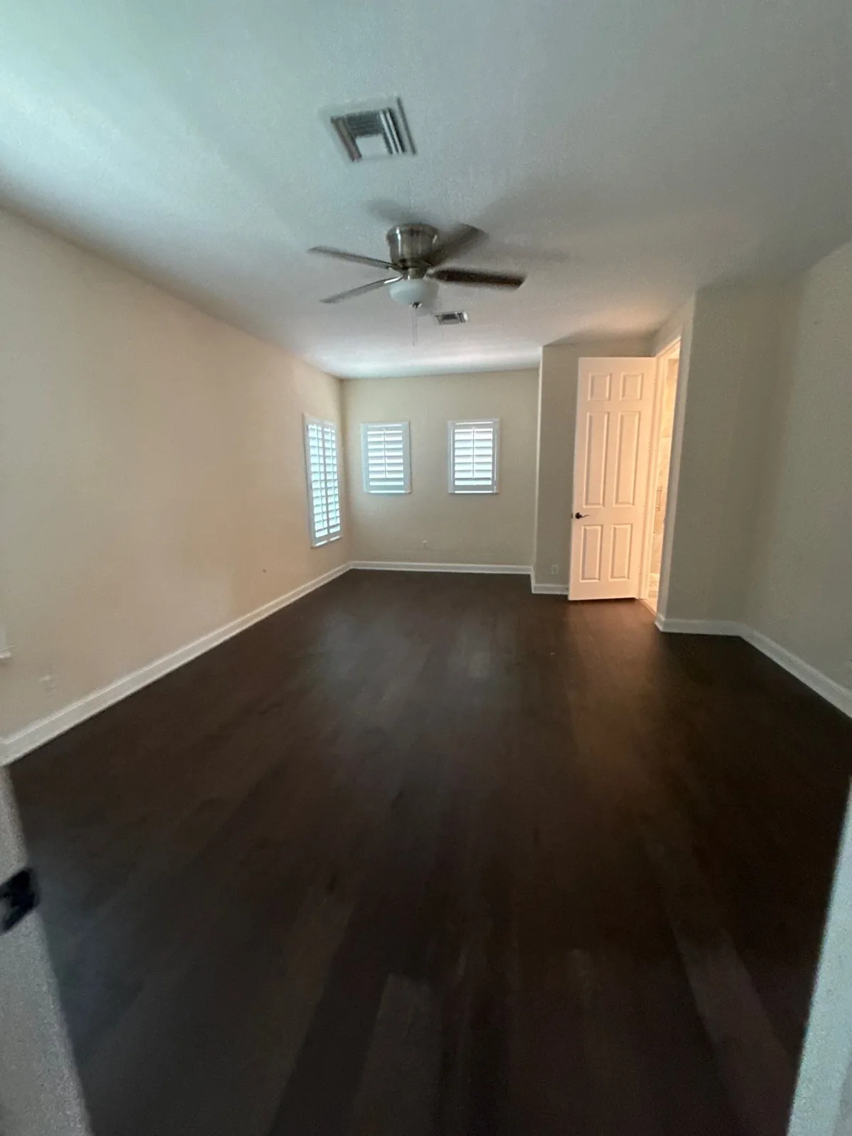 Interior Painting in North Palm Beach, Florida. Empty room with neutral painted walls, white six-panel door, plantation shutters, ceiling fan and dark wood flooring.