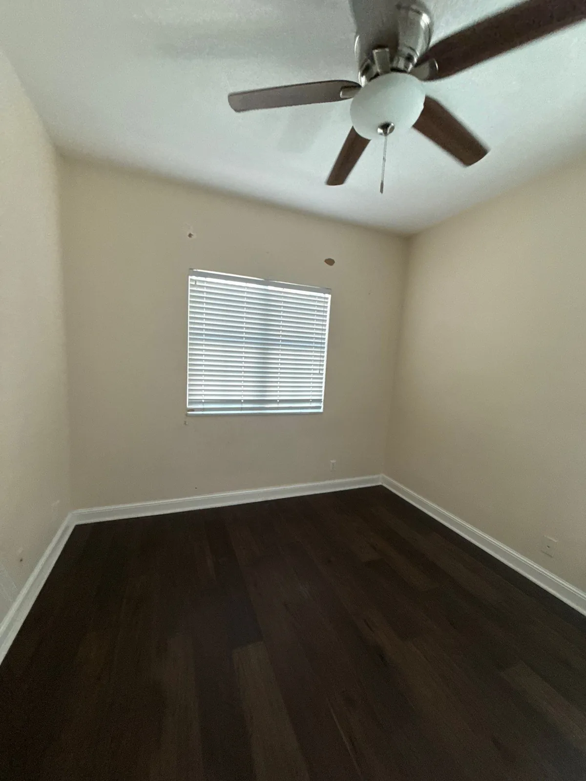 Interior Painting in North Palm Beach, Florida. Empty bedroom with beige walls, white baseboards, blinded window, ceiling fan and dark wood-look flooring.