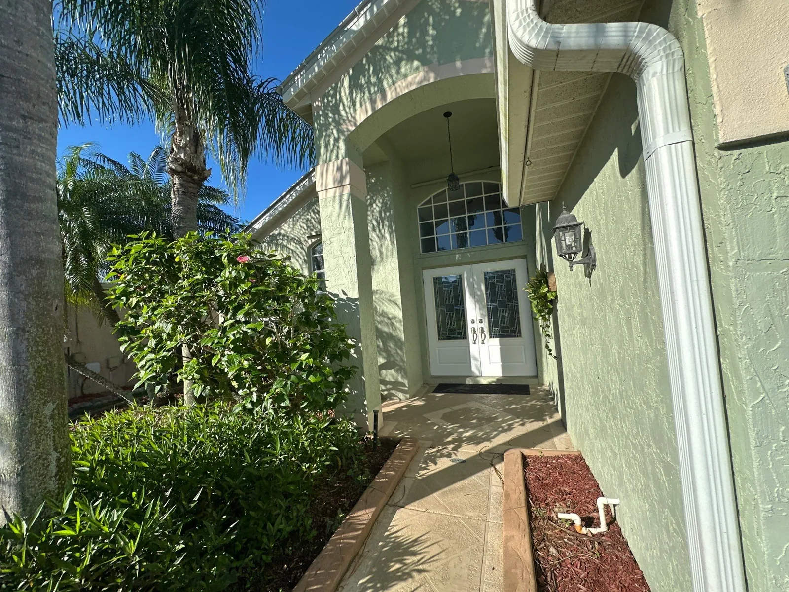Exterior Painting in North River Shores, Florida. Front entry of a pale green stucco home with arched portico, white double front door and white trim, landscaped walkway.