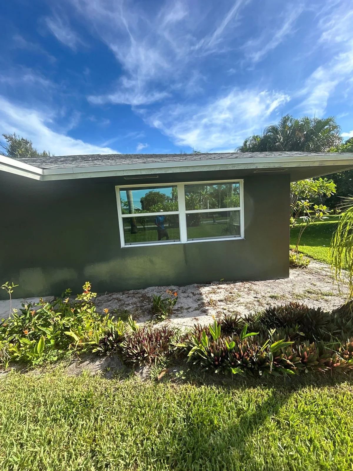 Exterior Painting in North River Shores, Florida. Corner of a single-storey house with dark green painted stucco walls and a white-trimmed window, under a clear blue sky.