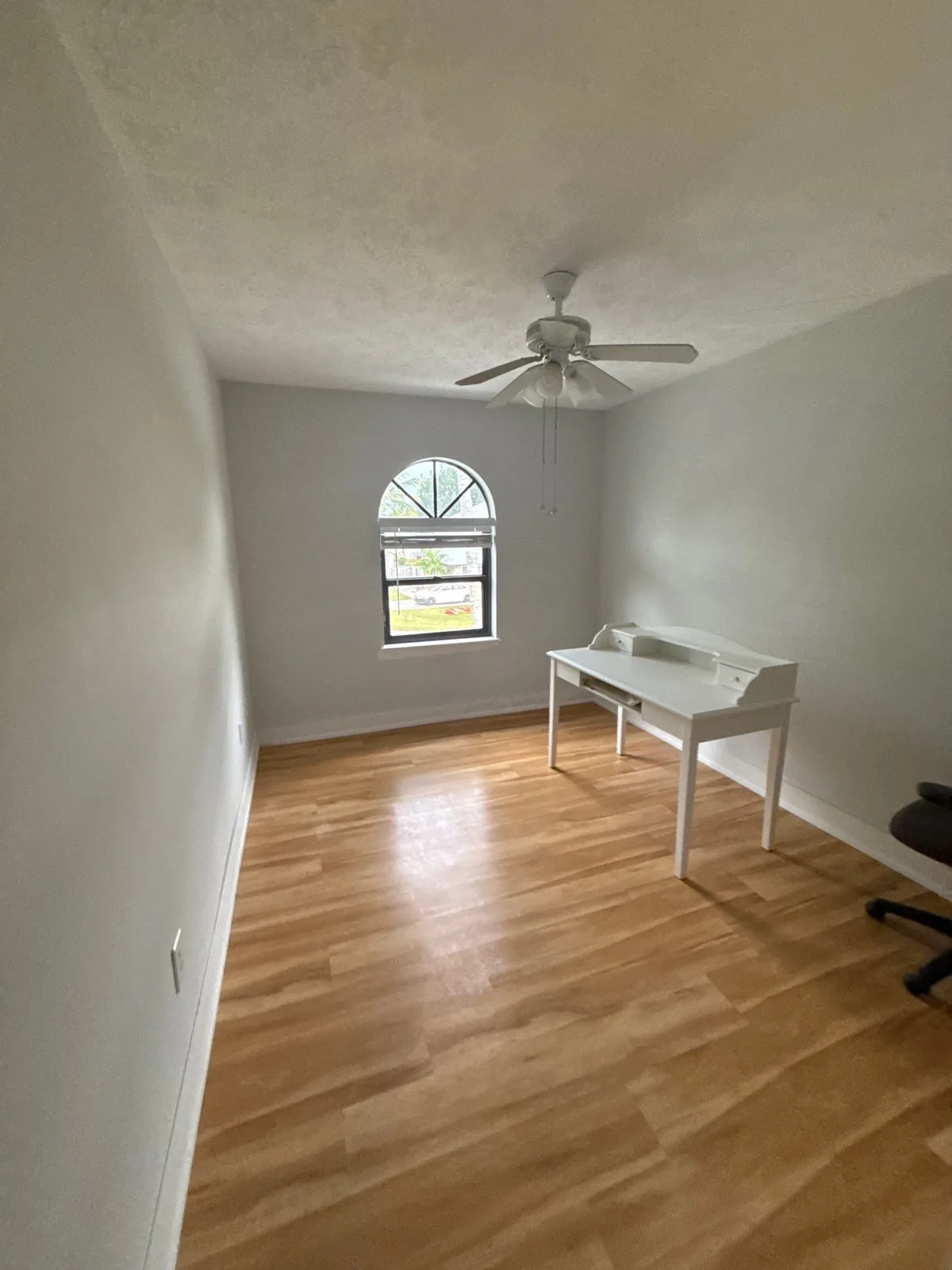 Interior Painting in North River Shores, Florida. Empty bedroom with grey painted walls, white baseboards, textured ceiling with fan, arched window and light wood-look flooring, with a small white desk.