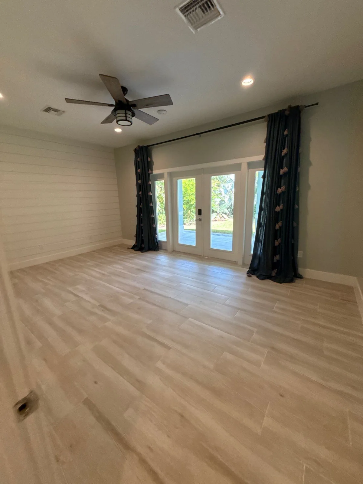 Interior Painting in North River Shores, Florida. Empty room with sage-grey walls, white trim, French doors flanked by dark curtains, ceiling fan and light wood-look floor.