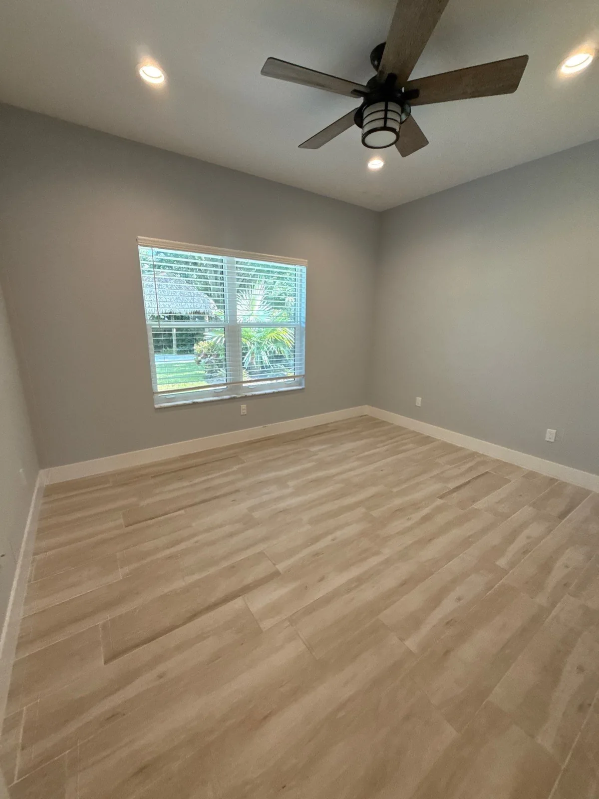 Interior Painting in North River Shores, Florida. Empty bedroom with grey walls, white window and baseboard trim, ceiling fan and light wood-look tile floor.