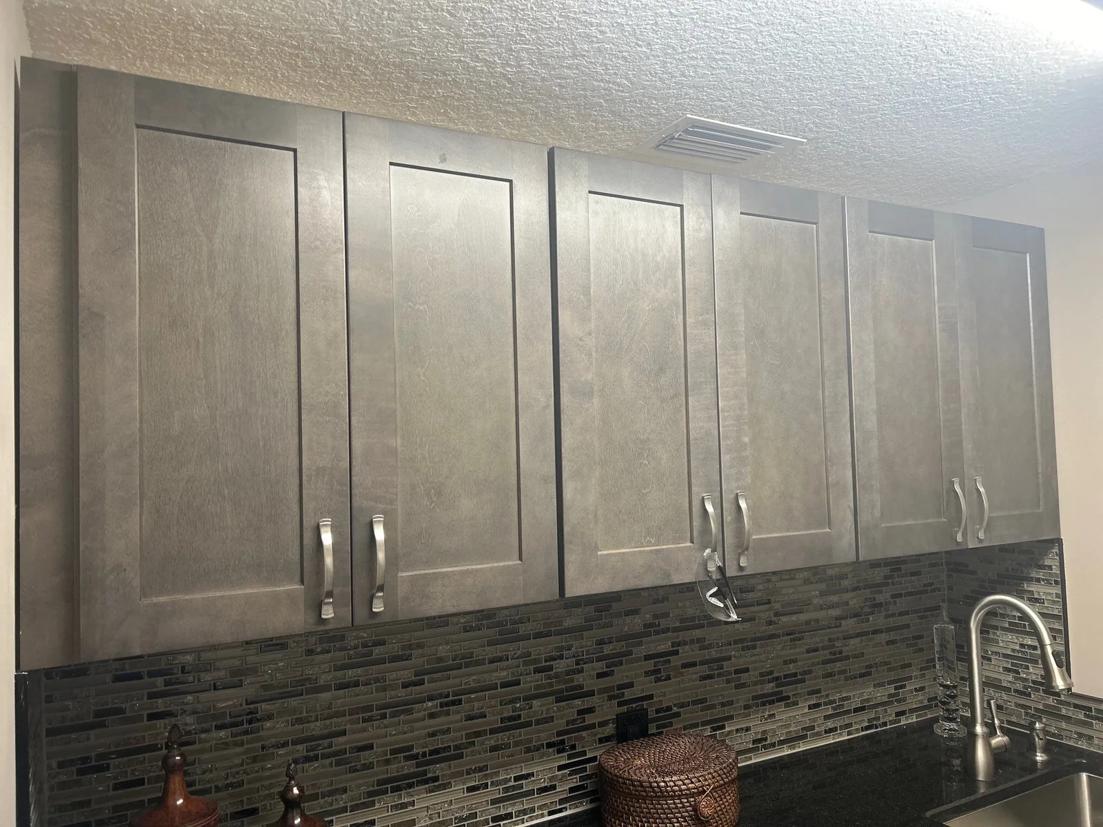 Cabinet Refinishing in Okeechobee County, Florida. Run of grey shaker-style upper cabinets with brushed metal bar pulls above a mosaic tile backsplash and dark speckled countertop.
