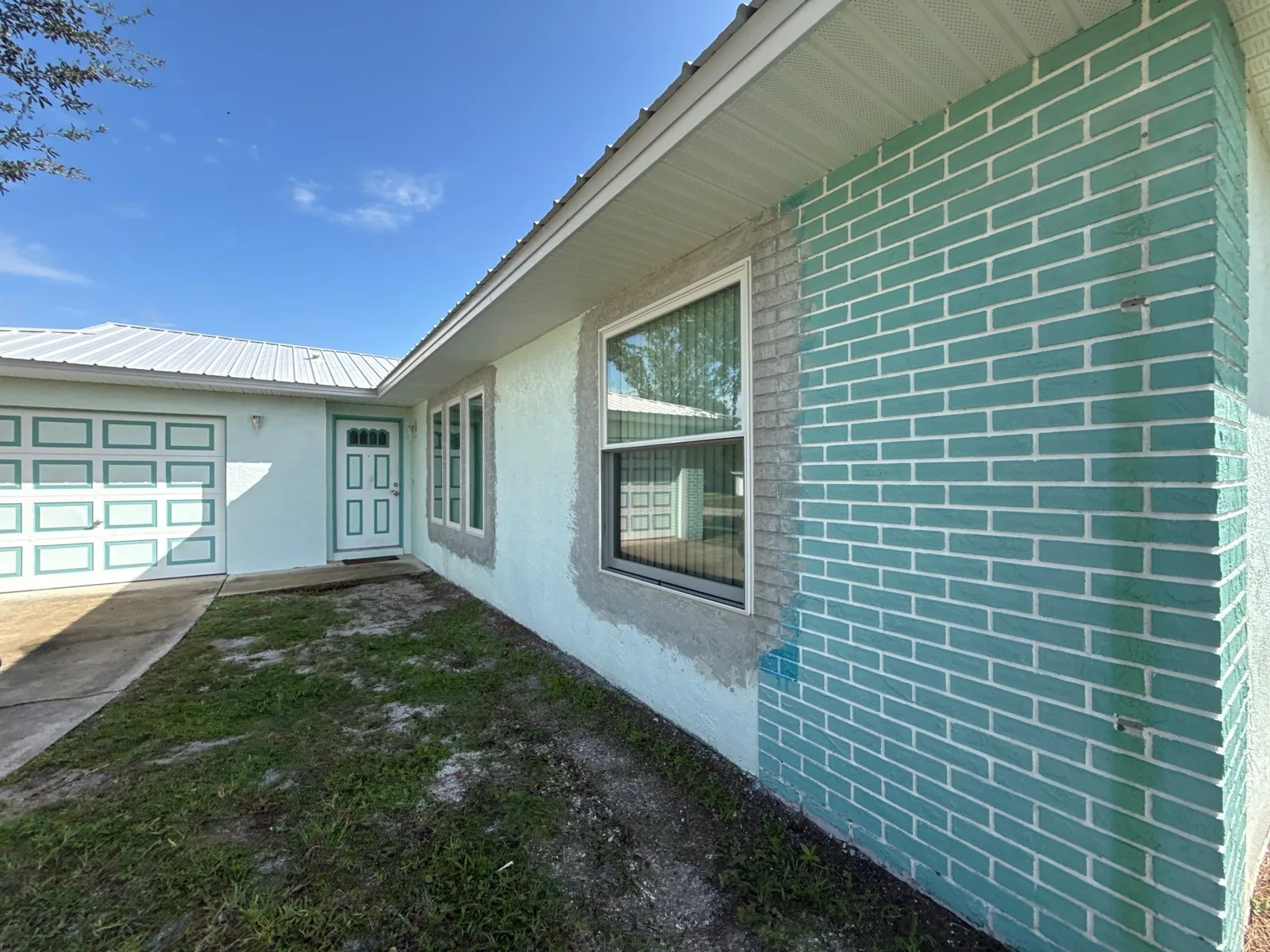 Exterior Painting in Palm Bay, Florida. Side elevation of a single-storey house with mint green painted brick, cream stucco, white trim, white window frame and white garage door.