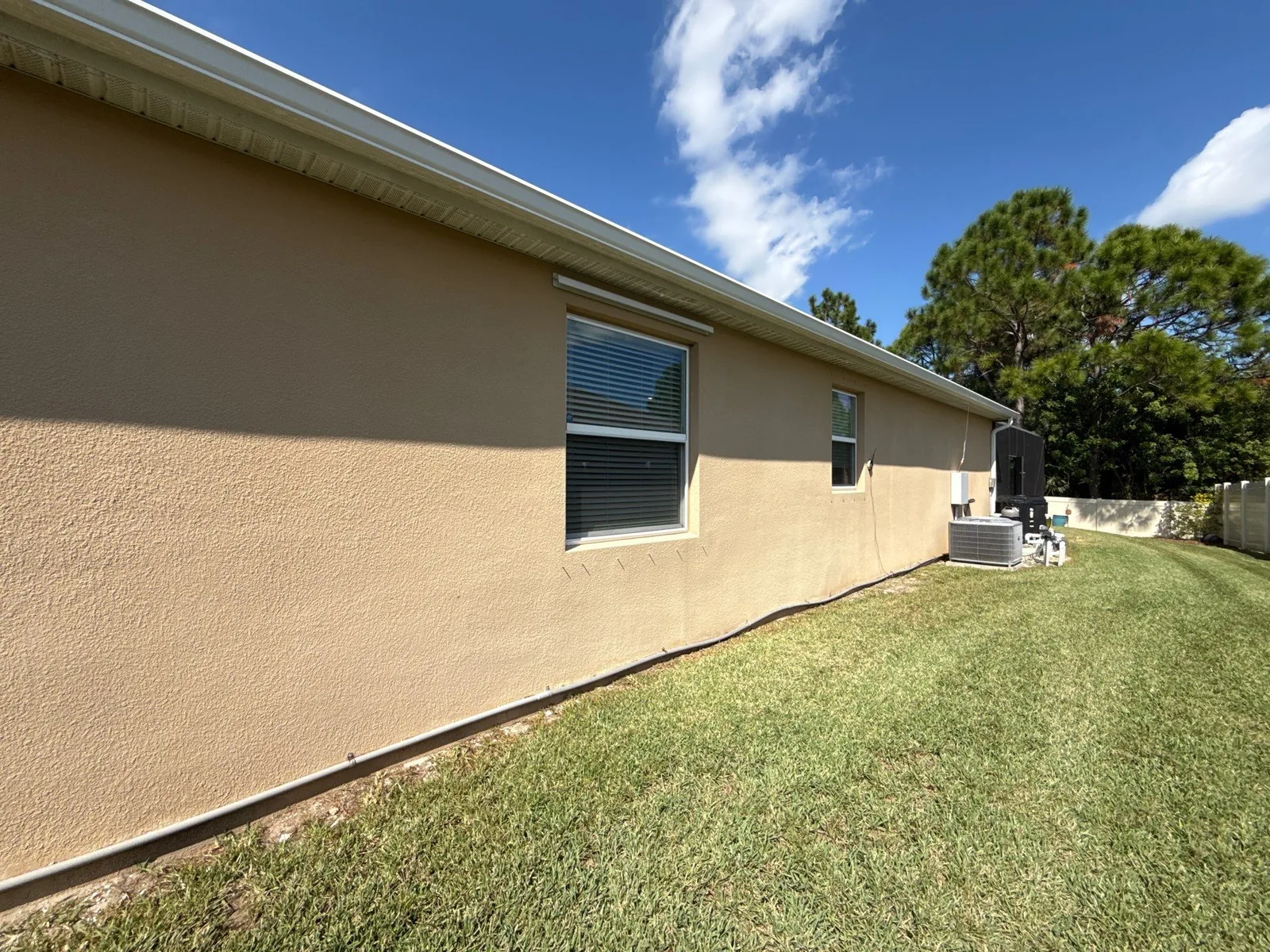 Exterior Painting in Palm Bay, Florida. Side elevation of a single-storey stucco house painted beige, with white fascia, two windows and a green lawn under blue sky.