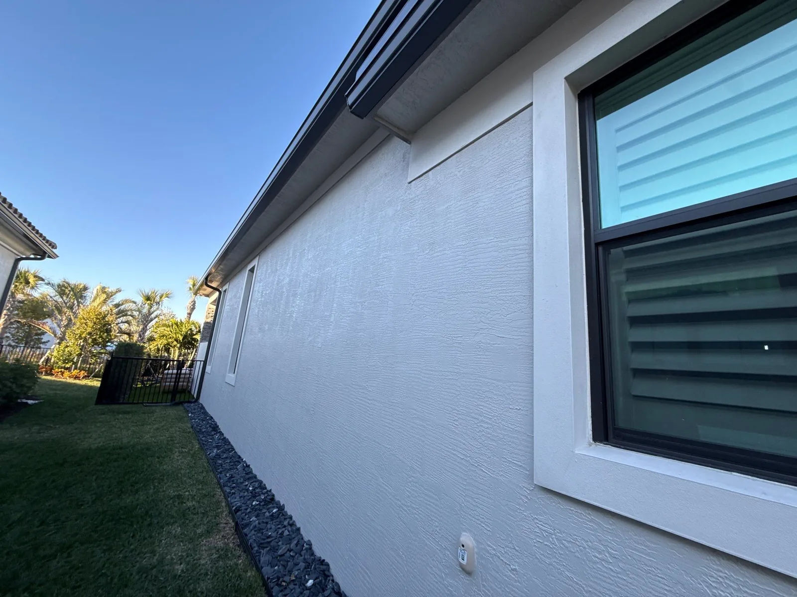 Exterior Painting in Palm Beach Gardens, Florida. Side elevation of a single-storey stucco home painted white with dark window frames, rock border and lawn under a clear blue sky.