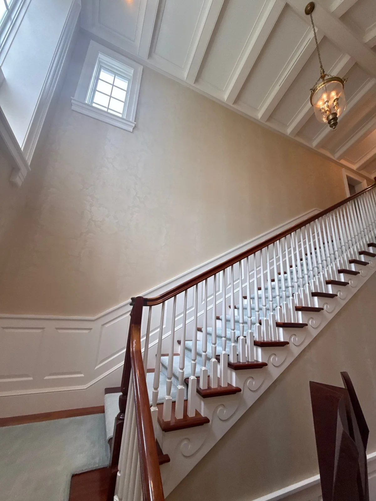 Interior Painting in Palm Beach, Florida. Staircase with white painted balusters and newel, wood handrail, white picture-frame wainscot, decorative scrollwork on the stringer, textured cream walls and a white beamed ceiling above.